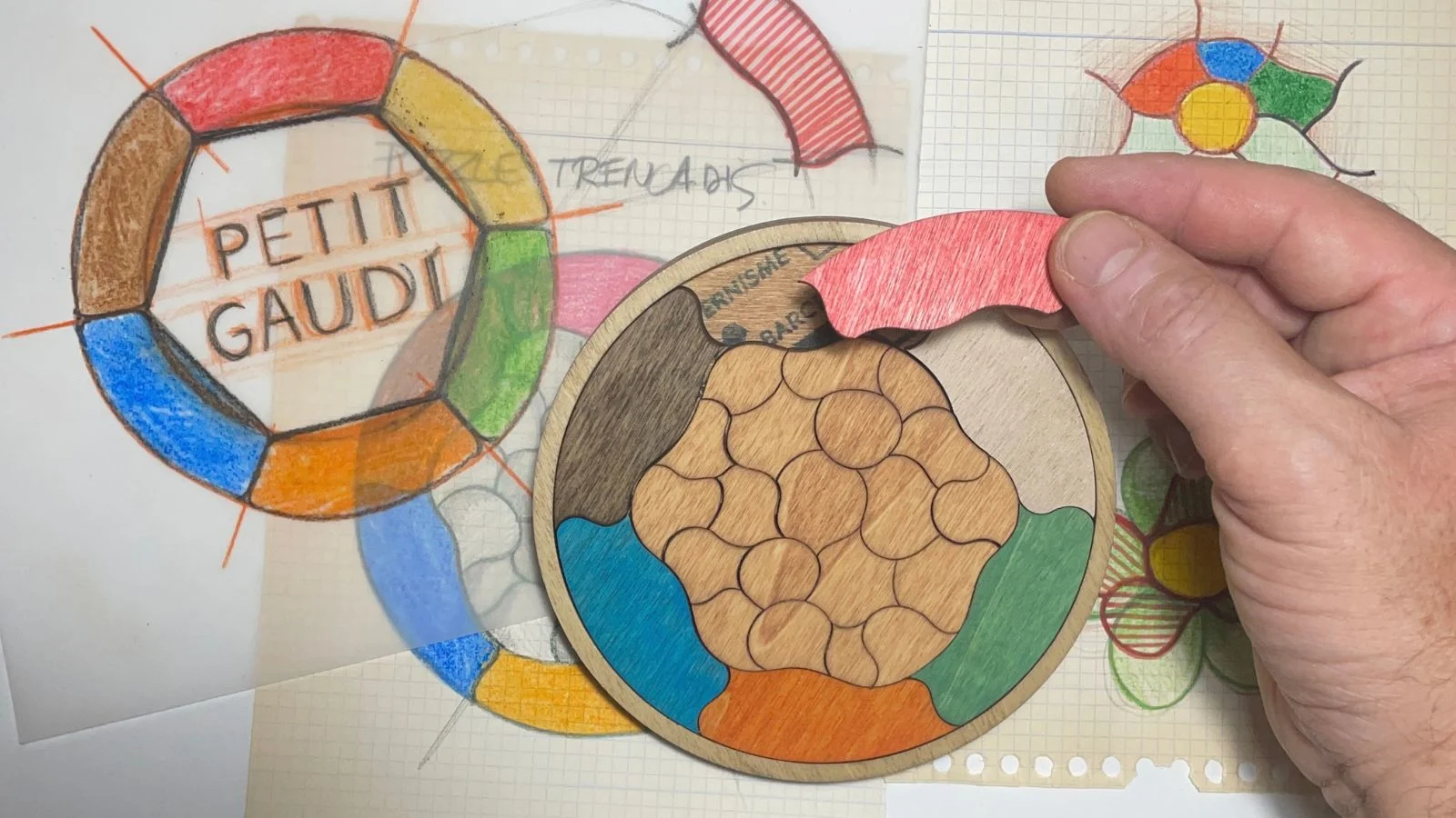 Boceto del logo de Petit Gaudí junto al Puzzle Trencadís de madera, mientras una mano retira una pieza roja.