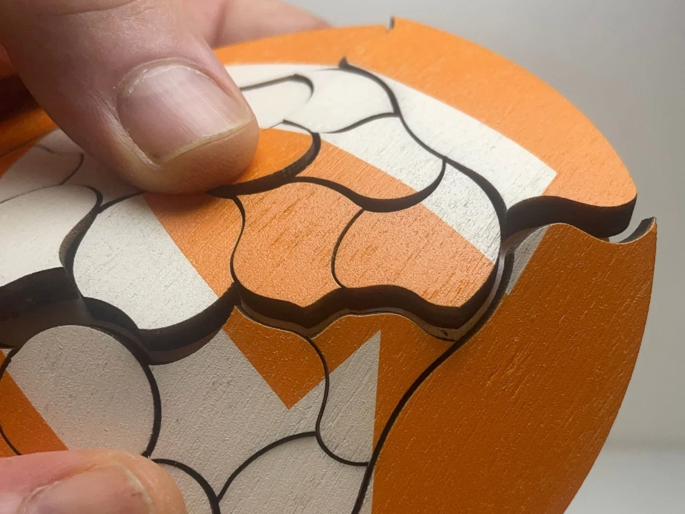 Una mano separa una pieza del Puzzle Trencadís de madera en naranja, blanco y negro.