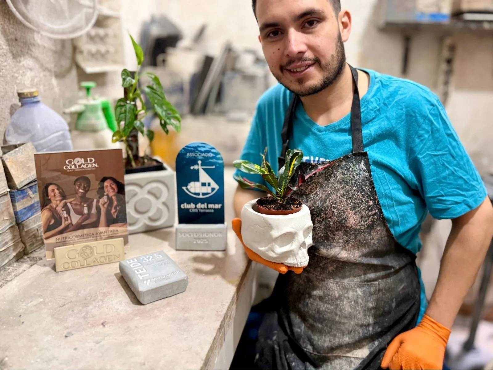 Carlos Baquero, diseñador y artesano de En Concreto Design Lab Carlos Baquero con camiseta azul y delantal, sosteniendo una maceta con una planta en su taller.